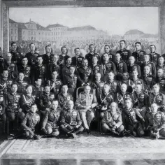 Grupa mężczyzn w mundurach wojskowych, w tym jeden w fotelu. W tle obraz przedstawiający budynki i tłum. bbwr.