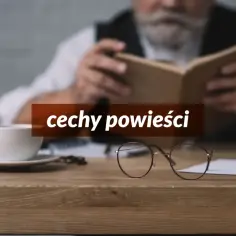 Starszy pan z siwą brodą czyta książkę. Na biurku stoi filiżanka kawy i okulary. Tekst na zdjęciu: "cechy powieści".
