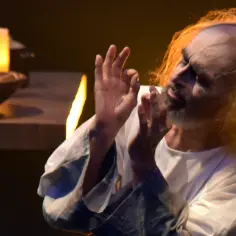 Aktor w białej szacie, z rozwianymi włosami, gestykuluje. Scena przypomina "Dziady" w reżyserii Rollisona.