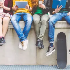 Grupa młodych ludzi, prawdopodobnie pokolenie Z, siedzi na murku. Każdy zajęty swoim urządzeniem lub książką. Obok deska do skateboardingu.