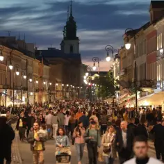 Tłum ludzi spaceruje wieczorem po oświetlonej ulicy. Widać kamienice, latarnie i kościelną wieżę. Każdy szuka czegoś dla siebie, jak oportunista na targu.
