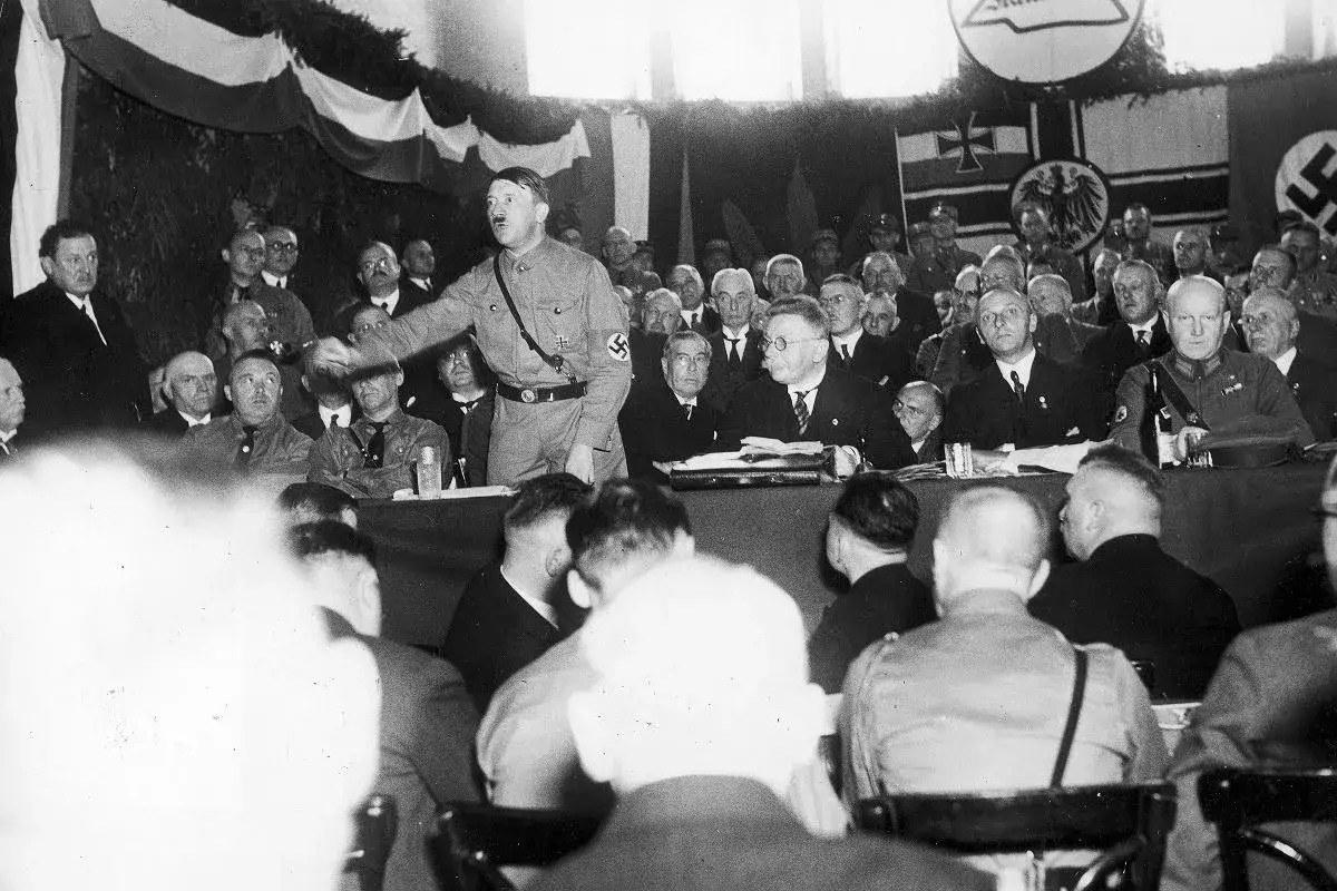 Adolf Hitler przemawia do tłumu, wyjaśniając, czym było NSDAP. W tle flagi i symbole partii.