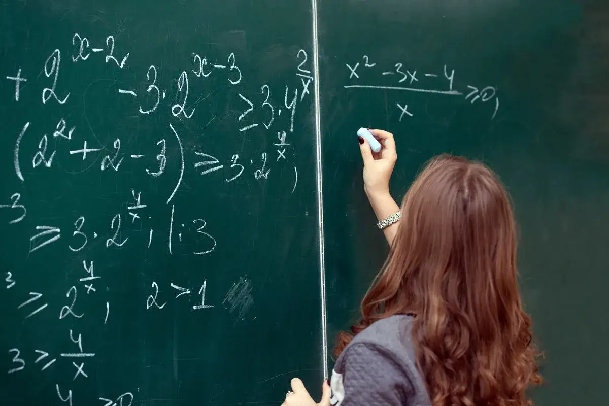 Dziewczyna pisze kredą równania na tablicy. Matematyczne zadania, pełne symboli i liczb, to przykład funkcji fatycznej w komunikacji – skupienie na samym akcie przekazywania informacji.