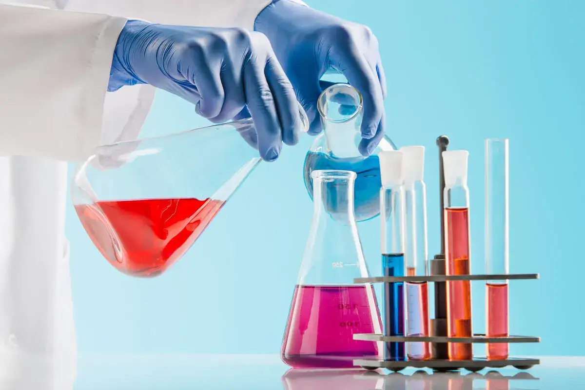 Laborant w niebieskich rękawiczkach przelewa czerwony płyn do kolby, dążąc do chemicznego puryzmu. Obok stoją probówki z kolorowymi substancjami.