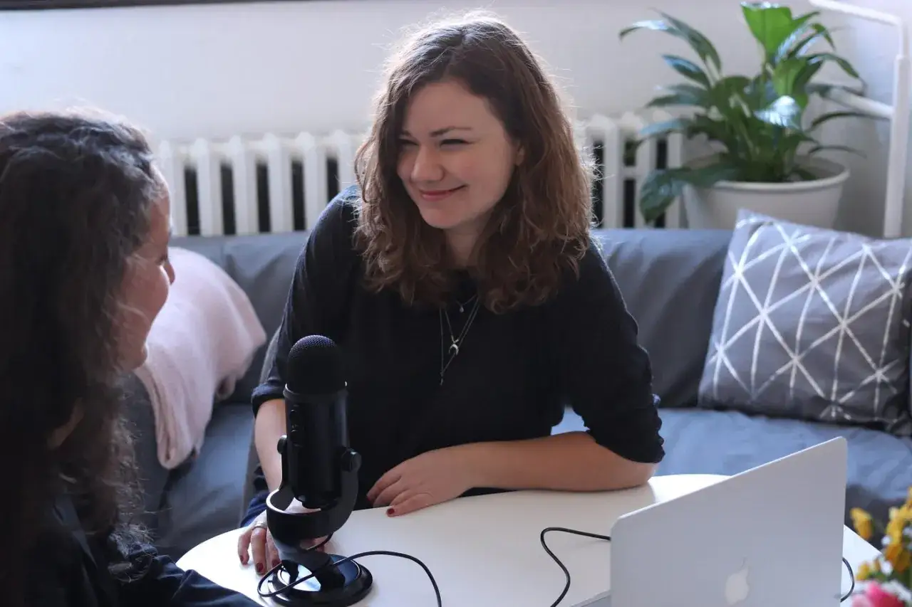 Dwie kobiety prowadzą wywiad przy mikrofonie i laptopie. Jedna z nich uśmiecha się, słuchając drugiej.