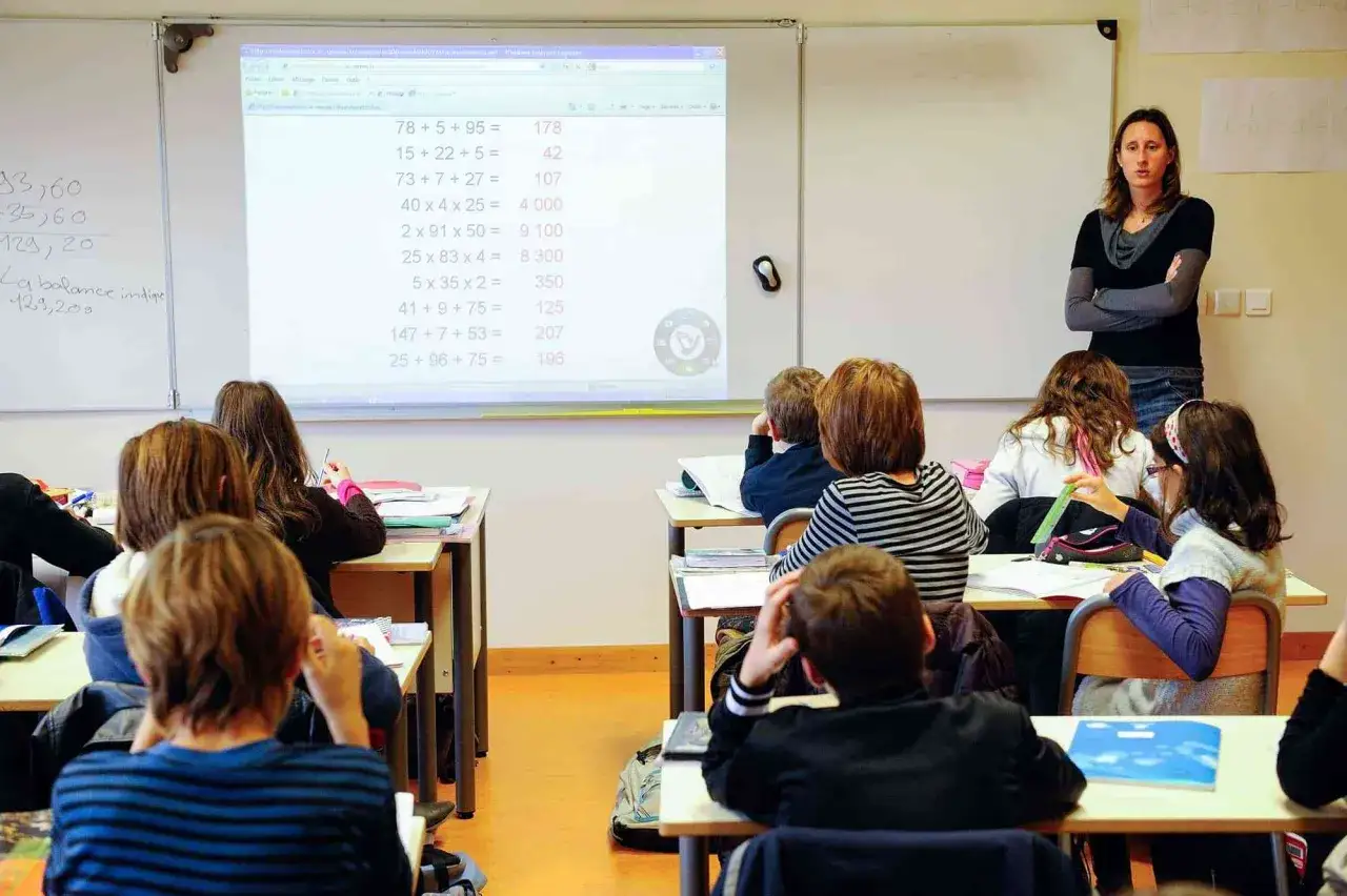 Nauczycielka tłumaczy zadania matematyczne. Uczennice i uczniowie uważnie słuchają. Widać feminatywy w nazwach zawodów, np. nauczycielka.