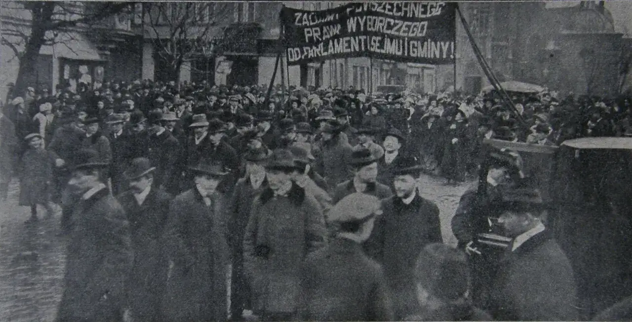 Tłum ludzi na ulicy, wśród nich sufrażystki, niesie transparent z hasłem o prawach wyborczych.
